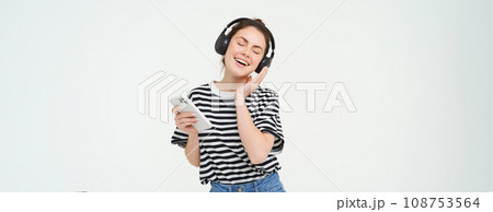 Young woman with smartphone listening to music, dancing to her favourite song in headphones, posing against white background Young woman with smartphone listening to music, dancing to her favourite song in headphones, posing against white background 108753564