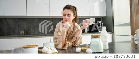 Attractive young cheerful girl baking at the kitchen, making dough, holding recipe book, having ideas Attractive young cheerful girl baking at the kitchen, making dough, holding recipe book, having ideas 108753631