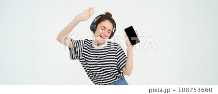 Portrait of stylish young woman dancing in wireless earphones, using headphones and mobile phone music app to listen to favourite songs, posing over white background 108753660
