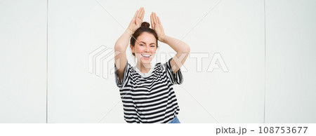Young woman laughing, showing animal floppy ears gesture with hands on top of head, smiling and looking happy, isolated over white background 108753677