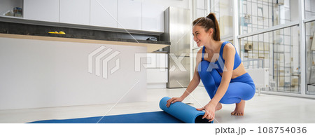 Health and sport concept. Young sportswoman at home, unwrapping her yoga mat, preparing living room for fitness, yoga workout 108754036