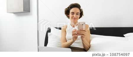 Work from home concept. Young woman sitting on bed, freelancing, working on laptop, using smartphone, looking at mobile phone with pleased smiling face Work from home concept. Young woman sitting on bed, freelancing, working on laptop, using smartphone, looking at mobile phone with pleased smiling face 108754097