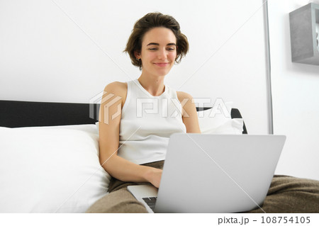 Portrait of smiling modern woman, sitting on her bed with laptop, looking happy, working from home, checking her emails on computer, browsing online shops Portrait of smiling modern woman, sitting on her bed with laptop, looking happy, working from home, checking her emails on computer, browsing online shops 108754105