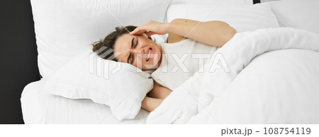Close up portrait of female model, lying in bed with headache, grimacing and frowning from painful migraine, has pain in head, feeling unwell 108754119