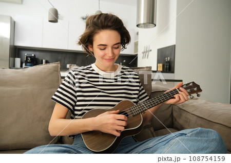 Portrait of young modern woman, student playing ukulele at home, sitting with small guitar, singing and feeling happy, sitting on sofa 108754159