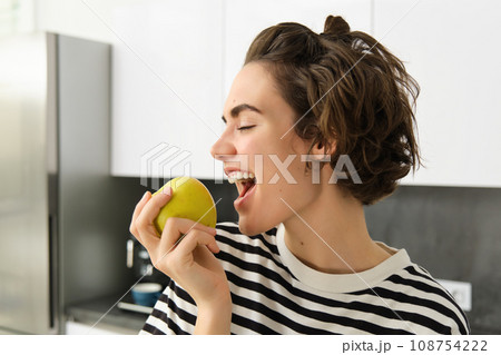 Close up portrait of young brunette woman biting an apple with pleasure, has pleased smile on her face, standing in the kitchen, having healthy snack for lunch, eating fruits 108754222