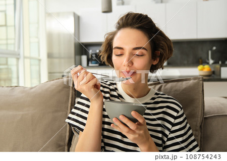 Portrait of young woman eating granola, bowl of cereals with milk, sitting on sofa and having her breakfast 108754243