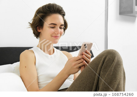 Cute young woman with mobile phone in hands, lying in bed in tank top, looking at smartphone with pleased smile 108754260