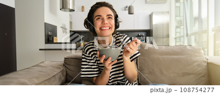 Portrait of woman watching tv in wireless headphones, looking at screen with interest and excitement, eating cereals in bowl, holding spoon, sitting on sofa in living room 108754277