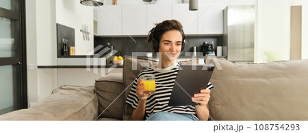 Portrait of woman in headphones, sitting on sofa with tablet and drinking orange juice, watching tv show on her gadget, using social media application, spending time at home 108754293