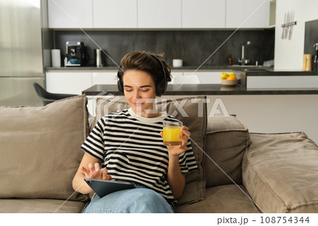 Close up portrait of modern girl, drinking orange juice, watching videos on digital tablet, sitting on sofa in living room Close up portrait of modern girl, drinking orange juice, watching videos on digital tablet, sitting on sofa in living room 108754344