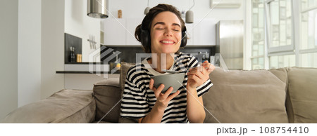 Portrait of woman laughing and smiling in headphones, eating cereals for breakfast and sitting on sofa in front of tv Portrait of woman laughing and smiling in headphones, eating cereals for breakfast and sitting on sofa in front of tv 108754410