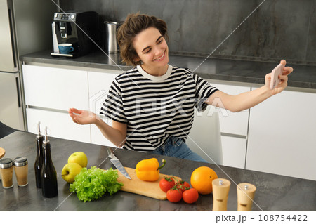 Portrait of beautiful young woman, food blogger recording video of her cooking in the kitchen, showing meal ingredients, preparing a meal on camera, using smartphone for live streaming Portrait of beautiful young woman, food blogger recording video of her cooking in the kitchen, showing meal ingredients, preparing a meal on camera, using smartphone for live streaming 108754422