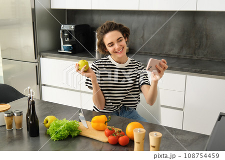 Smiling young woman, food blogger recording a video on smartphone with food, cooking and making selfie, taking pictures during meal preparation, sitting near vegetables and holding an apple 108754459