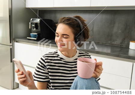 Close up portrait of young female student, drinking tea and listening to music in headphones, using smartphone in kitchen Close up portrait of young female student, drinking tea and listening to music in headphones, using smartphone in kitchen 108754488