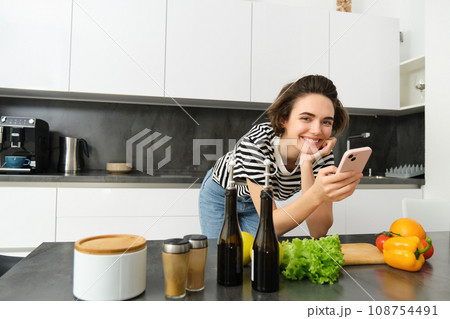 Portrait of woman, lifestyle blogger, posting picture of her cooking process on smartphone app, standing near vegetables and chopping board Portrait of woman, lifestyle blogger, posting picture of her cooking process on smartphone app, standing near vegetables and chopping board 108754491