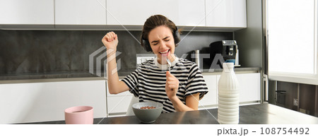 Carefree young woman, student in headphones, eating cereals for breakfast, listening music, singing and using soon as a microphone, having fun while having her meal in the morning Carefree young woman, student in headphones, eating cereals for breakfast, listening music, singing and using soon as a microphone, having fun while having her meal in the morning 108754492