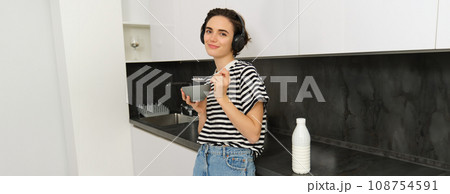 Portrait of modern woman eats breakfast, holds bowl and spoon, stands in kitchen, listens music in wireless headphones. Concept of lifestyle and home Portrait of modern woman eats breakfast, holds bowl and spoon, stands in kitchen, listens music in wireless headphones. Concept of lifestyle and home 108754591