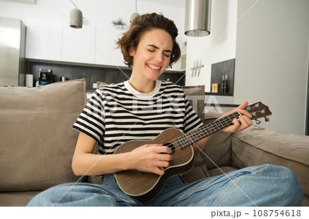 Portrait of young modern woman, student playing ukulele at home, sitting with small guitar, singing and feeling happy, sitting on sofa Portrait of young modern woman, student playing ukulele at home, sitting with small guitar, singing and feeling happy, sitting on sofa 108754618