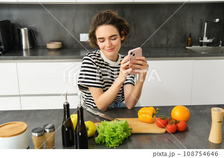 Portrait of young woman with smartphone, sitting in the kitchen and cooking salad, searching for healthy recipe online, has vegetables and chopping board on the table 108754646