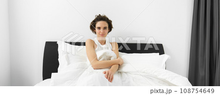 Image of young sensual woman with short hair, wearing tank top pyjama, lying in bed under white linen sheets, looking at camera, waking up in morning Image of young sensual woman with short hair, wearing tank top pyjama, lying in bed under white linen sheets, looking at camera, waking up in morning 108754649