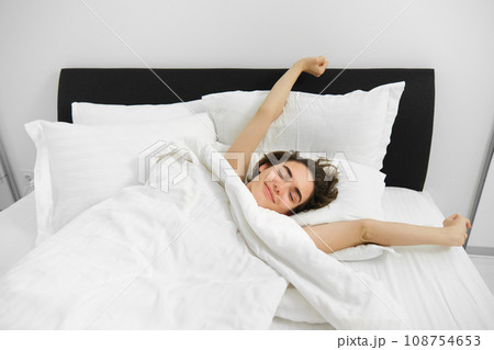 Portrait of happy, pleased young woman, stretching her arms with satisfied smile, lying in bed of soft pillow, waking up alone in bed, had good night sleep Portrait of happy, pleased young woman, stretching her arms with satisfied smile, lying in bed of soft pillow, waking up alone in bed, had good night sleep 108754653