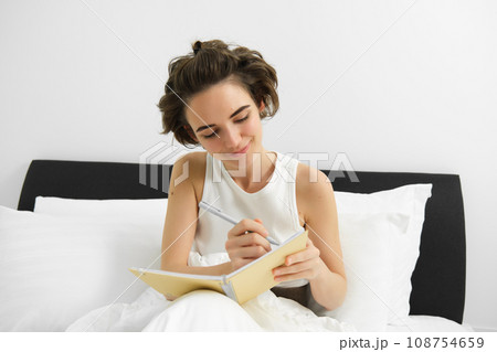 Image of mindful young woman sitting in bed, waking up and writing in journal, making notes, sharing her thoughts in diary 108754659