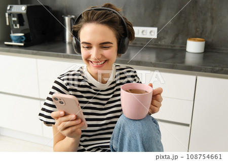 Close up portrait of young modern woman in earphones, listens music or podcast while drinking her morning tea, using smartphone, scrolling social media app Close up portrait of young modern woman in earphones, listens music or podcast while drinking her morning tea, using smartphone, scrolling social media app 108754661