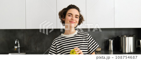 Portrait of beautiful, smiling young woman, holding an apple, eating fruit in the kitchen, looking happy. Concept of healthy diet and lifestyle Portrait of beautiful, smiling young woman, holding an apple, eating fruit in the kitchen, looking happy. Concept of healthy diet and lifestyle 108754687