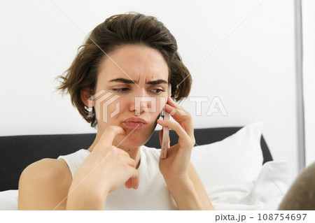 Close up portrait, woman makes a phone call with puzzled, frowning face, hearing complicated story over the telephone, talking to someone while resting in bed Close up portrait, woman makes a phone call with puzzled, frowning face, hearing complicated story over the telephone, talking to someone while resting in bed 108754697