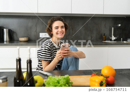 Portrait of young woman searching for cooking recipes online using smartphone, sitting near vegetables, salad ingredients and chopping board, smiling at camera Portrait of young woman searching for cooking recipes online using smartphone, sitting near vegetables, salad ingredients and chopping board, smiling at camera 108754711