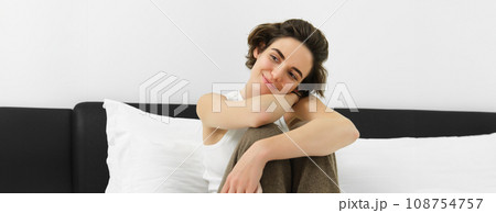 Young tender woman in her cosy pyjamas, sitting on bed in bedroom, smiling with sensual smile Young tender woman in her cosy pyjamas, sitting on bed in bedroom, smiling with sensual smile 108754757