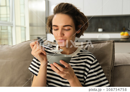 Portrait of young brunette woman eating on sofa, holding bowl and spoon, having cereals with milk for breakfast, enjoying her granola, spending time living room 108754758