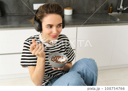 Young woman eating cereals in the morning, student having her breakfast, listening music or podcast in wireless headphones Young woman eating cereals in the morning, student having her breakfast, listening music or podcast in wireless headphones 108754766