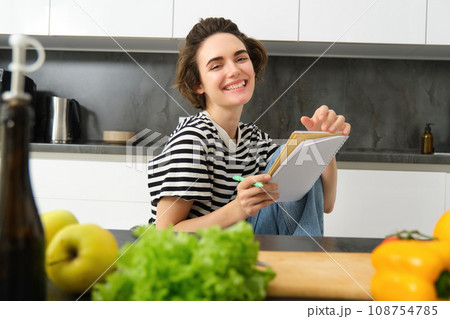 Portrait of young woman cooking, writing notes, grocery list in notebook, creating list of meals to cook through meal, sitting in kitchen near vegetables and chopping board Portrait of young woman cooking, writing notes, grocery list in notebook, creating list of meals to cook through meal, sitting in kitchen near vegetables and chopping board 108754785