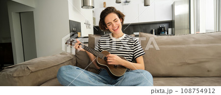 Portrait of cheerful young woman playing her ukulele, singing and laughing, sitting in living room at home Portrait of cheerful young woman playing her ukulele, singing and laughing, sitting in living room at home 108754912