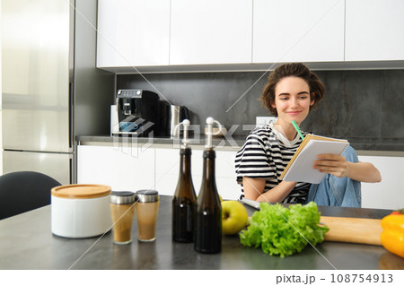 Portrait of young woman looking at cooking ingredients on kitchen counter and making notes, writing down recipes, thinking of meal for dinner, preparing vegetarian food Portrait of young woman looking at cooking ingredients on kitchen counter and making notes, writing down recipes, thinking of meal for dinner, preparing vegetarian food 108754913
