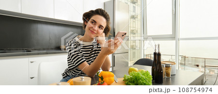 Portrait of brunette girl cooking food in the kitchen, searching recipes on social media app, holding mobile phone, standing near chopping board and vegetables, preparing healthy vegetarian meal 108754974