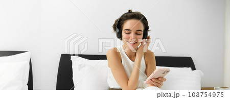 Portrait of smiling young woman in wireless headphones, sitting on bed with mobile phone, listening music in earphones 108754975