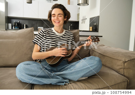 Portrait of cheerful young woman playing her ukulele, singing and laughing, sitting in living room at home Portrait of cheerful young woman playing her ukulele, singing and laughing, sitting in living room at home 108754994