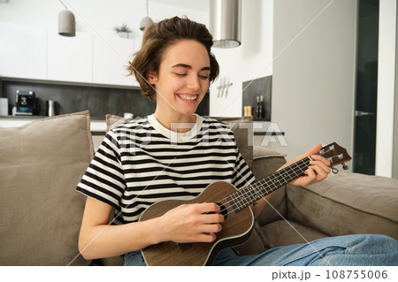 Portrait of young modern woman, musician playing ukulele at home, picking chords for new song, sitting on sofa in living room and smiling Portrait of young modern woman, musician playing ukulele at home, picking chords for new song, sitting on sofa in living room and smiling 108755006