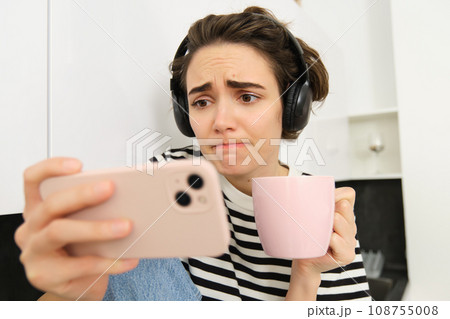 Close up portrait of young woman in headphones, watching something sad on smartphone application, frowning and looking with sympathy at mobile phone screen, drinking tea or coffee in the kitchen Close up portrait of young woman in headphones, watching something sad on smartphone application, frowning and looking with sympathy at mobile phone screen, drinking tea or coffee in the kitchen 108755008