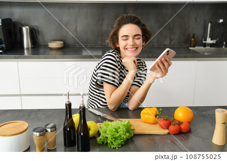 Happy smiling woman, sitting in the kitchen with smartphone, cooking salad, has chopping board, olive oil and vegetables on table 108755025