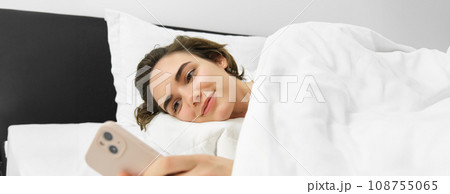Portrait of young woman lying in bed, going to sleep, waking up and checking her mobile phone, using smartphone in bedroom Portrait of young woman lying in bed, going to sleep, waking up and checking her mobile phone, using smartphone in bedroom 108755065