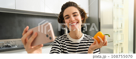 Close up portrait of cute brunette woman, taking selfie with yellow fresh pepper, recording video on smartphone, posing with vegetables in the kitchen, making cooking blog on social media app Close up portrait of cute brunette woman, taking selfie with yellow fresh pepper, recording video on smartphone, posing with vegetables in the kitchen, making cooking blog on social media app 108755070
