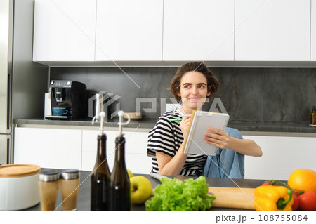 Portrait of young woman looking at cooking ingredients on kitchen counter and making notes, writing down recipes, thinking of meal for dinner, preparing vegetarian food 108755084