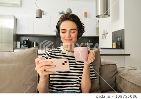 Young woman watching videos on smartphone app in headphones, drinking tea and sitting on sofa, spending comfort time at home Young woman watching videos on smartphone app in headphones, drinking tea and sitting on sofa, spending comfort time at home 108755086
