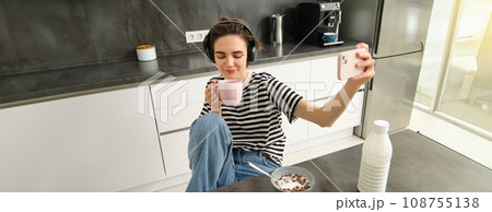 Portrait of young woman, content maker for social media, taking selfie while drinking tea and listening music in headphones, posing for photo, using smartphone to take pictures in the kitchen 108755138