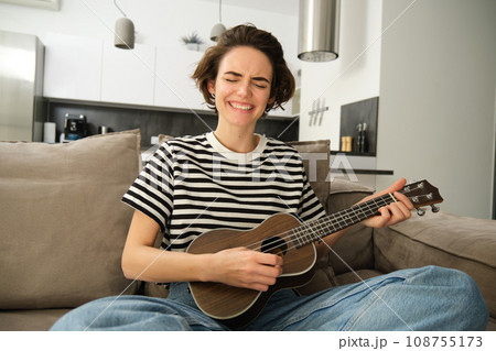 Passionate young woman, musician playing ukulele at home, singing with joy, sitting on couch in living room 108755173