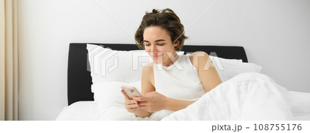 Portrait of young woman waking up in bed, using smartphone, turn off alarm on mobile phone, lying in her bedroom on white linen sheets 108755176